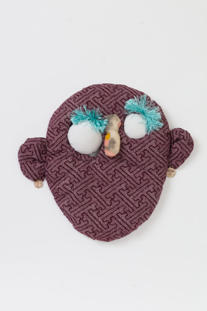 Et ansigt i lilla stof, med store hvide øjne og blå øjenvipper.