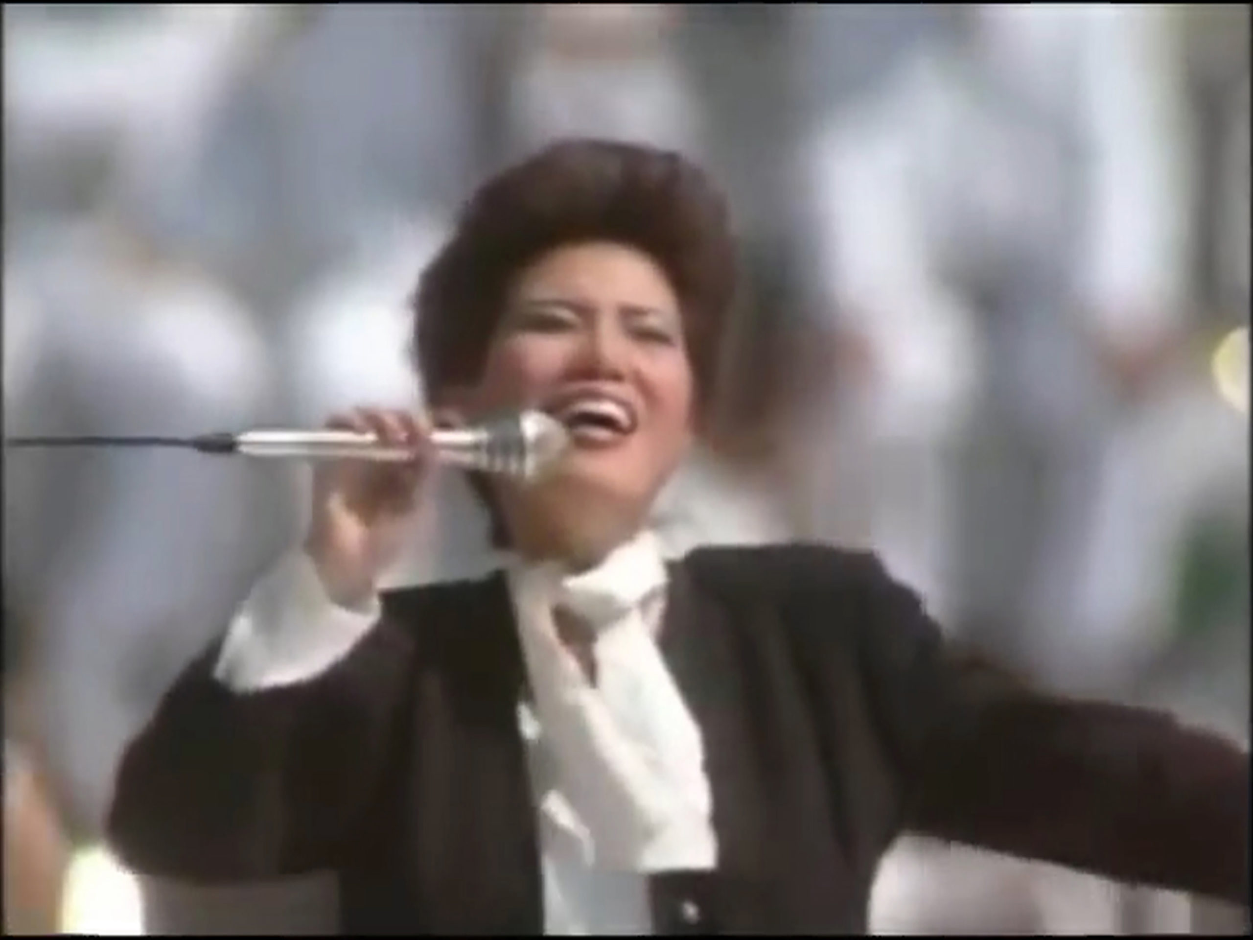 Woman with short hair sings energetically into a microphone, wearing a black jacket and white shirt with a bow, in front of a blurred background of people dressed in white.